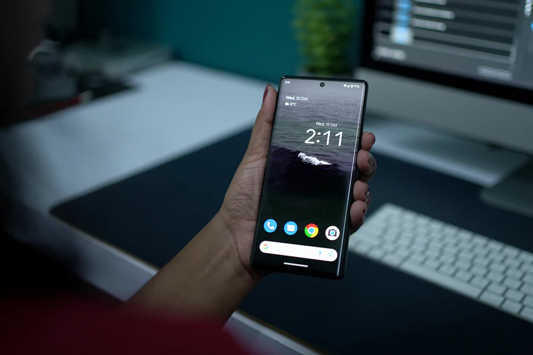 Mano sosteniendo un teléfono frente a un escritorio con computadora